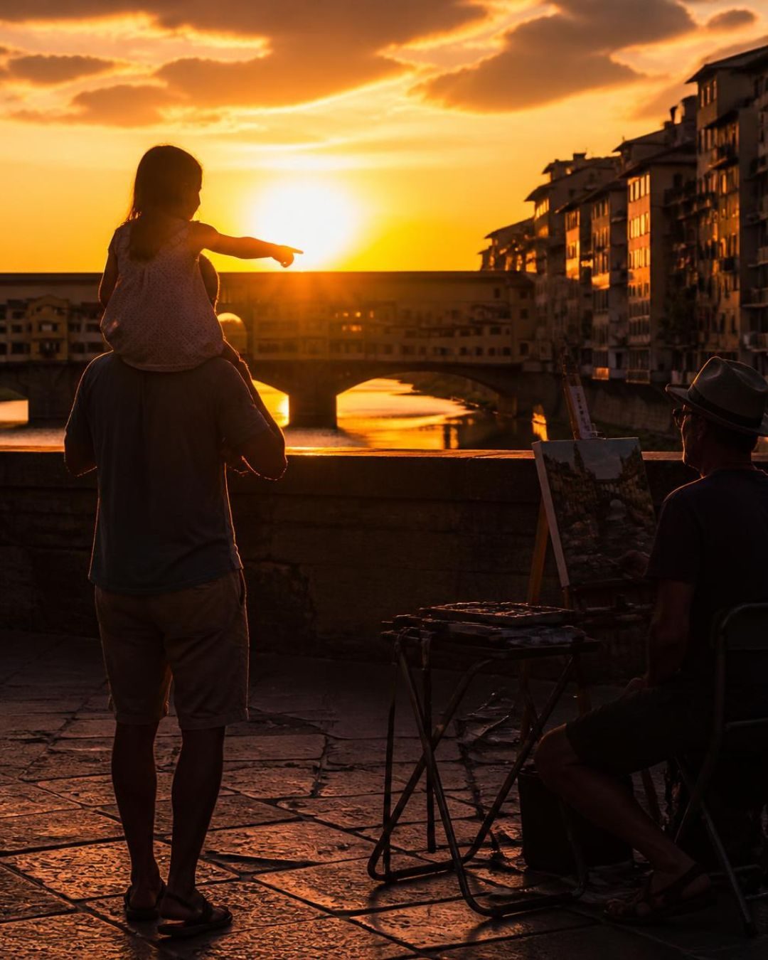Pai carregando a filha no ombro na Ponte Vecchio. Contraluz. Ela aponta para alguma coisa.
